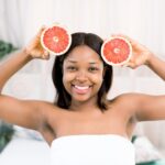 charming-young-african-girl-holding-slices-grapefruit-front-her-face-photo-smiling-african-american-woman-isolated-light-background-beauty-skin-care-concept_161094-2202